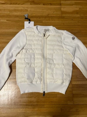 Vit cardiganjacka från Moncler - Stilren vit cardiganjacka från Moncler med quiltad front och stickade ärmar. Jackan har dragkedja framtill, ribbade muddar och Moncler-logga på ärmen. Perfekt för lager-på-lager och snygg till jeans eller kjol. För kvinnor i storlek 1/S. Kvitto dustbag och etiketter medföljer!