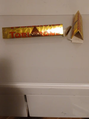 Guldig Toblerone-förpackning - Säljer en tom guldig Toblerone-förpackning i klassisk trekantig form. Förpackningen har röd och gul text och är tillverkad av papper. Perfekt för samlare eller pysselprojekt. Chokladen är uppäten och förpackningen är öppnad men annars okej skick.