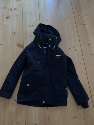 Svart jacka med huva från Dope - Snygg svart jacka från Dope med huva och vita detaljer. Jackan har flera fickor med tryckknappar, hög krage och justerbar huva.  Materialet är slitstarkt och vattenavvisande.