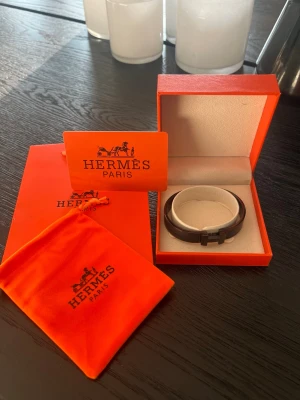 Svart armband från Hermès - Snyggt svart armband från Hermès med klassisk H-detalj framtill. Armbandet är tillverkat i metall och har en stilren, modern design. Levereras i original orange ask och tygpåse med Hermès-logga. Perfekt accessoar för att lyfta din outfit.