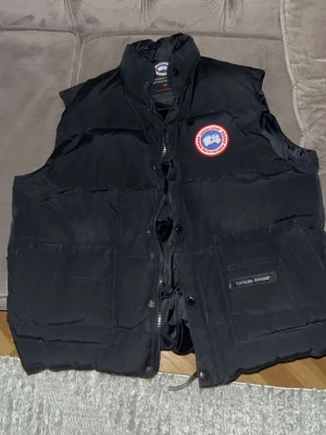 Svart dunväst från Canada Goose - Säljer en svart dunväst från Canada Goose med klassisk logotyp på bröstet. Västen har hög krage, dragkedja och knappar framtill samt stora fickor. Tillverkad i slitstarkt material och fylld med dun för extra värme. Perfekt för lager-på-lager och streetwear.