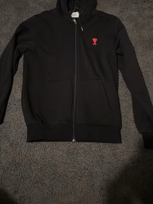 Svart hoodie med röd logga från Ami - Svart hoodie med dragkedja från Ami, prydd med en liten röd hjärtlogga på bröstet. Klassisk huva och två stora fickor framtill. Perfekt för en avslappnad och stilren look. Skriv om du kan ses i Stockholm 