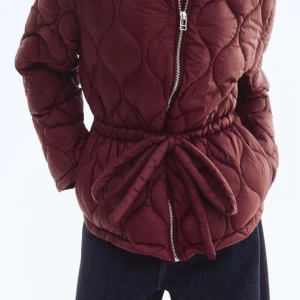 Vinröd quiltad pufferjacka från H&M - Säljer en vinröd pufferjacka från H&M med quiltat mönster, dragkedja framtill och matchande knytbälte i midjan. 