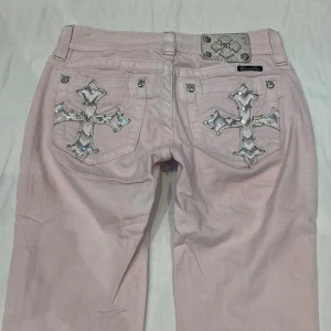 Ljusrosa jeans från Miss Me - Lågmidjade ljusrosa jeans med raka ben och snygga korsdetaljer i silver på bakfickorna. Jeansen har klassisk femficksdesign och cool metallpatch i midjan