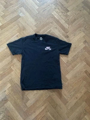 Svart Nike SB t-shirt med tryck - Cool svart t-shirt från Nike SB med liten logga på bröstet och stort grafiskt tryck i grönt och lila på ryggen. Klassisk passform och rund halsringning. Perfekt för dig som gillar streetwear och skatekultur.