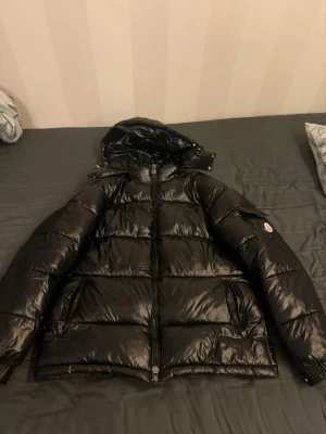 Svart Moncler dunjacka med huva - Snygg svart dunjacka från Moncler med glansig finish och huva. Jackan har dragkedja framtill, två fickor med dragkedja och en ficka med knapp på ärmen där Moncler-loggan sitter. Perfekt för kalla dagar och riktigt trendig look.