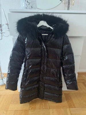 Glansig dunjacka med pälsluva - Jacka från LUXESWE med namn Luxegloss Long Black. Storlek XS, men passar mig som vanligtvis har S. Hemsidans beskrivning  är ”vattentät dunjacka med äkta pälskrage”. Nypris 3399:- (men nedsatt på deras hemsida just nu till 2889:-). Köpt 2022 men endast använd 2-3 gånger. Försluts med dragkedja + knappar. Två fickor med dragkedja. Rekommenderar att gå in på deras hemsida luxeswe.com för ännu bättre bilder!