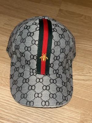 Gucci keps med monogram och rand - Snygg keps från Gucci i grått med klassiskt GG-monogram över hela ytan. Framtill löper en grön och röd rand med en gul broderad insekt. Kepsen har böjd skärm och justerbar rem baktill. Materialet är canvasliknande tyg. OBS FAKE