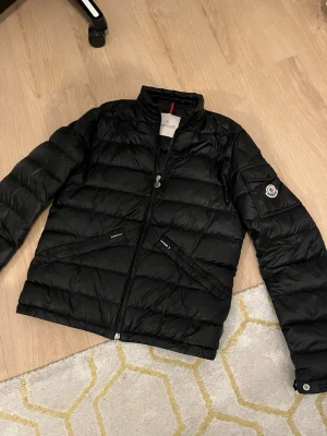 Svart dunjacka från Moncler - Snygg svart dunjacka från Moncler med klassisk quiltad design och hög krage. Jackan har två dragkedjeförsedda fickor framtill och en ficka med knapp på ärmen där Monclers logga är broderad. Perfekt för kalla vinterdagar och riktigt stilren.