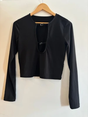 Svart långärmad croppad topp - Trendig svart långärmad topp med croppad passform och unik öppen detalj framtill som hålls ihop med två spännen. Perfekt för dig som vill sticka ut med en edgy stil. Mjuk och stretchig känsla.