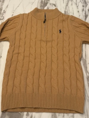Beige kabelstickad tröja Polo Ralph Lauren - Kabelstickad tröja i beige från Polo Ralph Lauren med halv dragkedja och ribbade muddar. Tröjan har en klassisk polokrage och den ikoniska mörkblå logotypen broderad på bröstet. Perfekt för lager på lager och snygg till jeans.