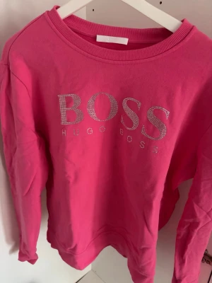 Rosa sweatshirt från Hugo Boss - Snygg rosa sweatshirt från Hugo Boss med stora glittriga bokstäver på bröstet. Tröjan har rund halsringning, långa ärmar och ribbade muddar. Tillverkad i mjukt bomullsmaterial som känns skönt mot huden. Perfekt för en trendig och avslappnad stil.