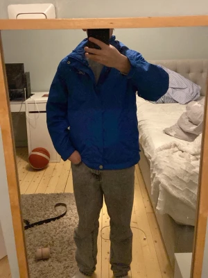 Blå vindjacka från FiveSeasons - Snygg blå vindjacka från FiveSeasons med huva och dragkedja framtill. Huvan sitter fast med tryckknappar så går att ta av. Jackan har två sidofickor, resår vid ärmsluten och tryckknappar vid halsen. Perfekt för blåsiga dagar och enkel att matcha med olika outfits.