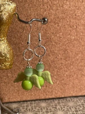 Gröna örhängen änglar  - Unika handgjorda örhängen med gröna pärlor och ljusgröna vingar i plast. Örhängena har silvriga krokar och ringar, och pärlorna är i två olika nyanser av grönt. Perfekt för dig som vill ha något lekfullt och färgglatt. Ej nickelfria 