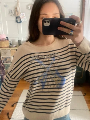 Zadi Voltaire Randig stickad tröja med gitarrmotiv - Stickad tröja i ljusbeige med mörkblå ränder och coolt tryck av två blå gitarrer på bröstet. Rund halsringning och knappar på axeln. Perfekt för dig som gillar rockig stil och unika detaljer.