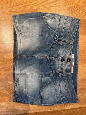 Blå jeanskjol med dubbla knappar - Säljer en blå jeanskjol i kort modell med dubbla knappar framtill och slitna detaljer. Kjolen har fram- och bakfickor samt bälteshällor. Perfekt för dig som gillar en avslappnad och trendig denimlook.