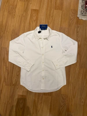 Vit skjorta från Polo Ralph Lauren - Klassisk vit skjorta från Polo Ralph Lauren med lång ärm och knappar framtill. Skjortan har den ikoniska broderade loggan på bröstet och är tillverkad i mjuk bomull. Perfekt för en clean och stilren look.