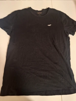 Svart t-shirt från Hollister - Klassisk svart t-shirt från Hollister med rund halsringning och liten vit logga på bröstet. Tillverkad i mjuk bomull för en bekväm känsla. Perfekt basplagg till garderoben och enkel att matcha med allt. Någon fläck under ena armen.
