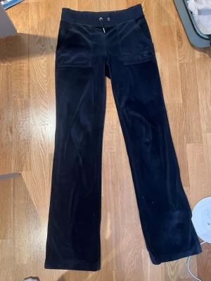 Svarta velourbyxor Juicy Couture - Mjukisbyxor i svart velour från Juicy Couture med raka ben, bred resår i midjan och snörning framtill. Byxorna har stora fickor fram och bak med knappdetaljer. Perfekta för en avslappnad och trendig look. Ett litet hål längs ner på benet som knappt syns 