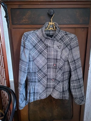 Grå rutig kavaj med knappar - Snygg grå kavaj från Zara med rutigt mönster och hög krage. Jackan har dekorativa knappar framtill och markerad midja med veckade detaljer på sidorna. Perfekt för dig som gillar en unik och trendig stil. (möts upp i Stockholm) 