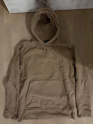 Beige/brun hoodie med ficka från J.lindeberg  - Säljer en beige hoodie från J.lindeberg med stor känguruficka framtill och justerbar huva med snören. Hoodien har en avslappnad passform och är tillverkad i mjukt bomullsmaterial. Perfekt för chill dagar och enkel att matcha med jeans eller sweatpants.