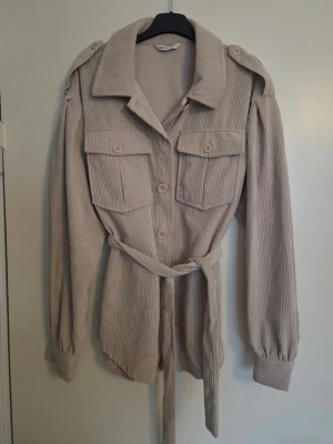 Beige overshirt i manchester - Stilren beige overshirt från Only i mjuk manchester. Modellen har stora bröstfickor, axelklaffar, knappar framtill och ett matchande knytskärp i midjan. Långa ärmar med manschett och avslappnad passform. Perfekt lager på lager-plagg.