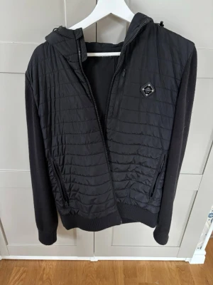 Svart cardigan från J.Lindeberg  - Svart jacka med quiltad framsida och rygg, stickade ärmar och huva. Jackan har dragkedja framtill, två fickor med dragkedja och en liten bröstficka. Perfekt för kyliga dagar och har en sportig vibe.