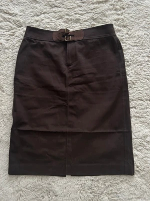 Brun kjol med spänne - Mörkbrun kjol med dubbla spännen i midjan. Rak modell som slutar ovanför knäet, perfekt för en stilren look. Materialet känns stabilt och passar till många olika outfits. Storlek 4, sitter ungefär som en M
