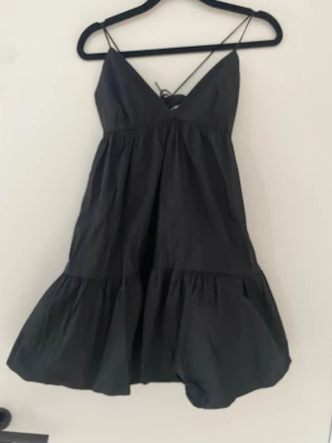 Svart kort klänning med tunna band - Säljer en svart kort klänning med tunna axelband och v-ringning. Klänningen har volang nedtill och snörning i ryggen för en snygg passform. Perfekt för sommardagar eller utekvällar. Materialet känns lätt och luftigt.
