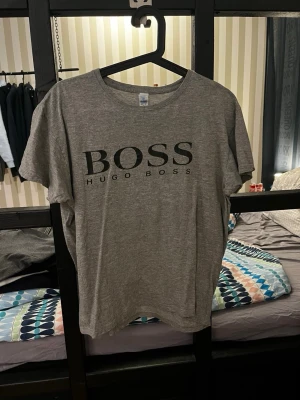 Hugo Boss t-shirt - Snygg grå t-shirt från Hugo Boss med stor svart logga på bröstet. Klassisk rund halsringning och korta ärmar. Perfekt för en stilren och avslappnad look.