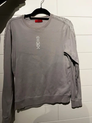 Hugo sweatshirt - Snygg grå sweatshirt från Hugo med vita detaljer på axlarna och ett diskret tryck på bröstet. Tröjan har rund halsringning och långa ärmar. Perfekt för en avslappnad och trendig stil.