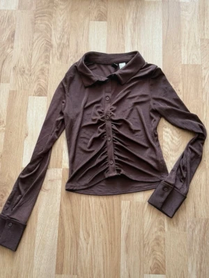 Brun skir blus med rynkade detaljer - Trendig brun skir blus med långa ärmar och knappar framtill. Skjortan har en snygg rynkning längs knappraden och klassisk krage. Perfekt för dig som gillar en unik twist på vardagslooken.