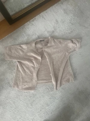 Beige kortärmad kofta - Snygg beige kofta med korta ärmar och rund hals. Koftan har en rak passform och knäppning framtill. Perfekt att slänga över en topp för en avslappnad look. Materialet känns mjukt och bekvämt. Dock saknar koftan några knappar!