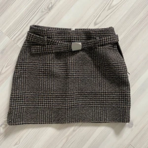 Rutigt minikjol med bälte - Snygg minikjol i svartvitt rutigt mönster med tillhörande bälte och silverfärgat spänne. Kjolen har en rak passform och är gjord i ett tjockare tyg som ger en stilren look. Perfekt för att lyfta vilken outfit som helst.