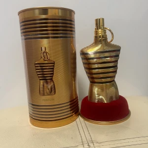 Jean Paul Gaultier parfym Elixir - Exklusiv parfym från Jean Paul Gaultier i en unik guldfärgad flaska formad som en manlig torso med horisontella soppränder. Kommer i matchande guldfärgad metalltub med samma design. Flaskan står på en rund röd sammetsbas. Perfekt för dig som gillar lyxig design.