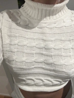 Vit kabelstickad croppad polotröja - Trendig vit kabelstickad croppad polotröja med höghals och långa ärmar. Snygg struktur med breda kabelstickade detaljer över hela tröjan. Perfekt för lager på lager och att styla till jeans eller kjol.