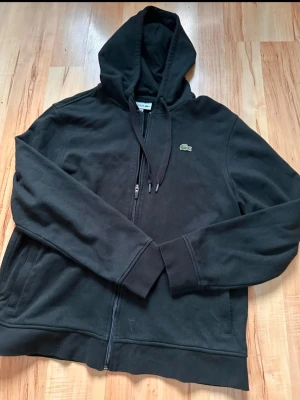Svart zip hoodie från Lacoste - Säljer en svart hoodie från Lacoste med dragkedja framtill och klassisk huva med dragsko. Ikoniska krokodilloggan på bröstet. Hoodien har långa ärmar, ribbade muddar och två fickor framtill.
