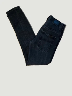 Svarta slim jeans från Nudie Jeans - Svarta/grå Nudie Jeans i storlek W30 L32 i en stilren slim fit-modell. Högkvalitativ denim med snygg lätt tvätt och naturligt slitage som ger ett tidlöst uttryck. Byxorna är i fint använt skick utan större skador. Observera att modellen upplevs något liten i storleken, passar bäst dig som normalt har W29 och gillar en tajtare passform. Perfekta både till vardag och mer uppklädda tillfällen.
