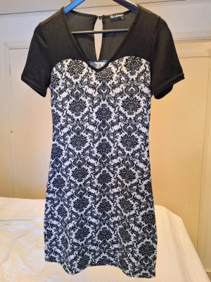 Svartvit abstrakt mönstrad miniklänning - Romantisk, klassisk & stilren - Kort klänning från Indulgence med svartvitt barockinspirerat mönster. Svart överdel med v-ringning och korta ärmar. Klänningen har en liten öppning med knapp i nacken och är tillverkad i supermjuk ullblandning. Storlek M/L, men mer som en medium utifrån mått. Stretchigt stickat tyg. Bystomkrets: ca 82 cm Längd från axel: ca 88 cm