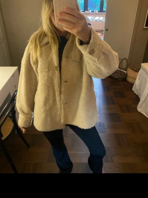 Beige teddyjacka med fickor - Mysig beige teddyjacka med oversized passform och stora bröstfickor. Jackan har krage och knäppning framtill med knappar. Perfekt för dig som gillar en avslappnad och trendig stil. Materialet är fluffigt och ger en riktigt skön känsla.