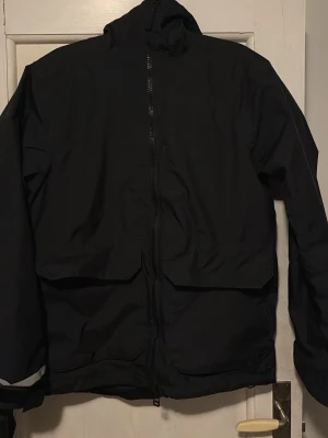 Svart vindjacka från Helly Hansen - Snygg svart vindjacka från Helly Hansen med hög krage och hel dragkedja framtill. Jackan har två stora fickor med lock och reflexdetalj på ena ärmen. Materialet är tunt och vindtätt, perfekt för blåsiga dagar.