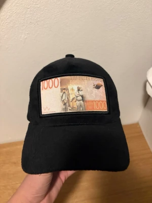 Svart keps med 1000-kronors patch - Cool svart keps med mesh på sidorna och en unik patch framtill som föreställer en svensk 1000-kronorssedel. Perfekt för dig som vill sticka ut och ha något annorlunda på huvudet. Justerbar baktill för bästa passform.