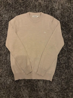Beige stickad tröja från J.Lindeberg - Snygg beige stickad tröja från J.Lindeberg med diskret broderad logga på bröstet. Tröjan har rund halsringning, långa ärmar och ribbade muddar vid ärmslut och nederkant. Perfekt för en stilren och enkel look.