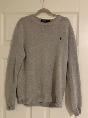 Grå stickad tröja från Polo Ralph Lauren - Klassisk grå stickad tröja från Polo Ralph Lauren med rund halsringning och diskret broderad logga i mörkblått på bröstet. Tröjan har ribbstickade muddar vid ärmslut och nederkant. Perfekt för lager-på-lager-stil.