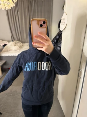 Mörkblå HOODRICH hoodie - Snygg mörkblå hoodie från HOODRICH med stor logga i vitt och blått på bröstet. Klassisk känguruficka framtill och justerbar huva med snören. Perfekt för en chill och street-inspirerad look. Mjuk och skön bomullskänsla.