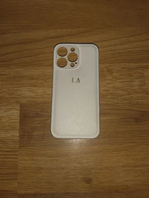 Vitt mobilskal med initialer i guld för iphone 13 pro - Snyggt vitt mobilskal med struktur och guldfärgade initialer 'I.A' på baksidan. Skalet har stora utskärningar för kameror och är gjort i ett material som liknar konstläder. Passar dig som vill ha en stilren och personlig look till mobilen.