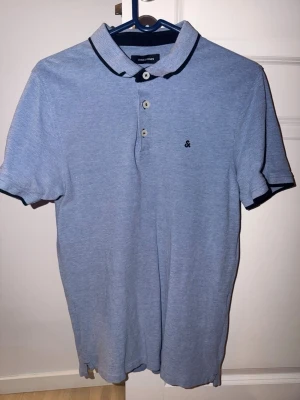 Blå piké från Jack & Jones - Snygg ljusblå pikétröja från Jack & Jones med mörkblå detaljer på krage och ärmslut. Klassisk krage, tre knappar framtill och liten broderad logga på bröstet. Perfekt till jeans eller shorts. Passar dig som gillar lite tightare passform.