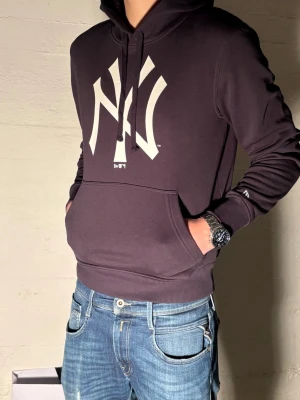 Svart New York Yankees hoodie - Snygg svart hoodie från New Era med stor vit New York Yankees-logga på bröstet. Klassisk känguruficka framtill, ribbade muddar och justerbar huva med snören. Perfekt för dig som gillar streetwear och sportig stil.
