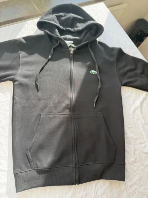 Svart zip hoodie från Lacoste - Säljer en svart hoodie från Lacoste med dragkedja, huva med snören och klassisk krokodil-logga på bröstet. Hoodien har känguruficka framtill och ribbade muddar vid ärmslut och nederkant. Perfekt för en chill och stilren look.