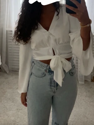 Vit knytblus med v-ringning - Trendig vit blus med v-ringning och knytning framtill. Blusen har långa, vida ärmar och är gjord i ett lätt, flowy material som ger en luftig känsla. Perfekt att styla med jeans eller kjol för en chic look.